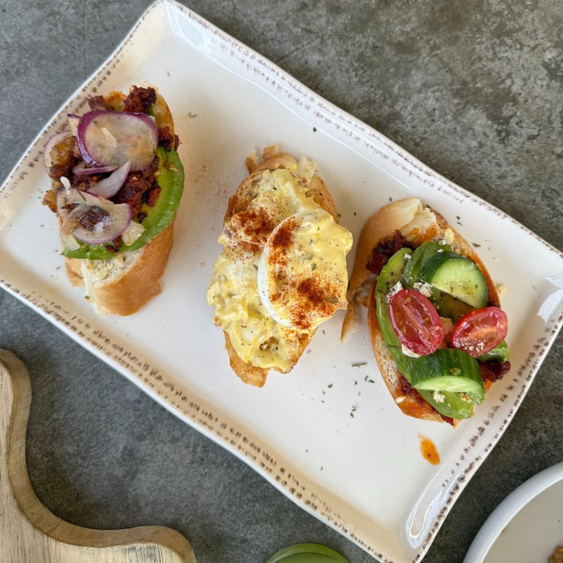 Three gourmet Sage Social Eatery bruschettas on a white rectangular plate, featuring various fresh toppings.