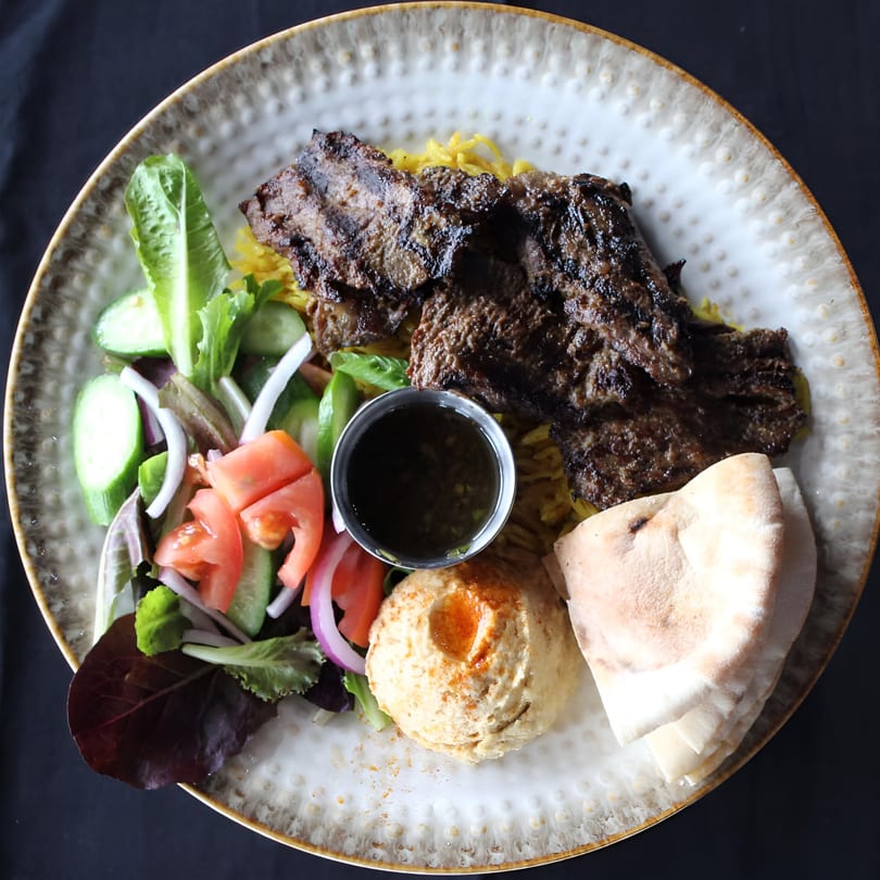 Grilled meat with yellow rice, salad, hummus, pita bread, and dipping sauce on a plate.
